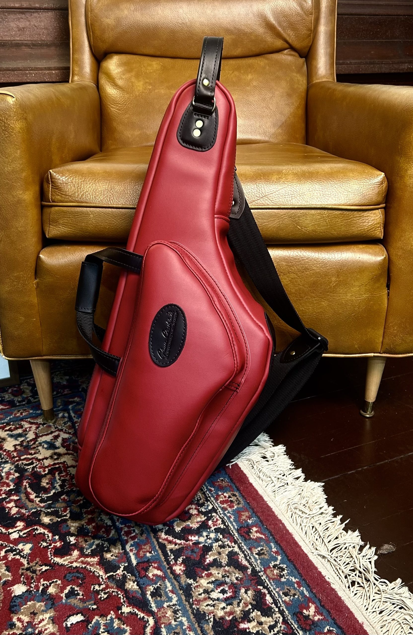 A red leather alto saxophone case is shown standing on its end on an oriental rug in front of a blonde-leather chair